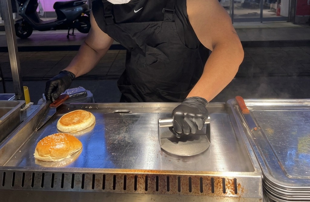 A person using a heavy metal burger press to firmly flatten a raw ball of ground beef onto a hot griddle.