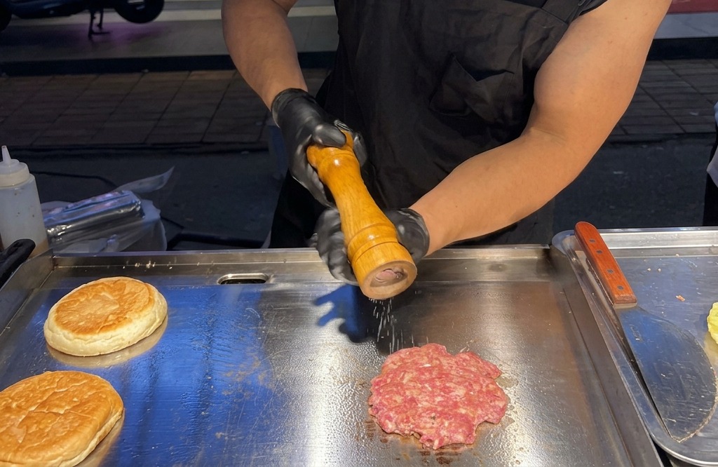 Grinding fresh sea salt and black pepper from a wooden mill onto a sizzling smashed beef patty on a griddle.