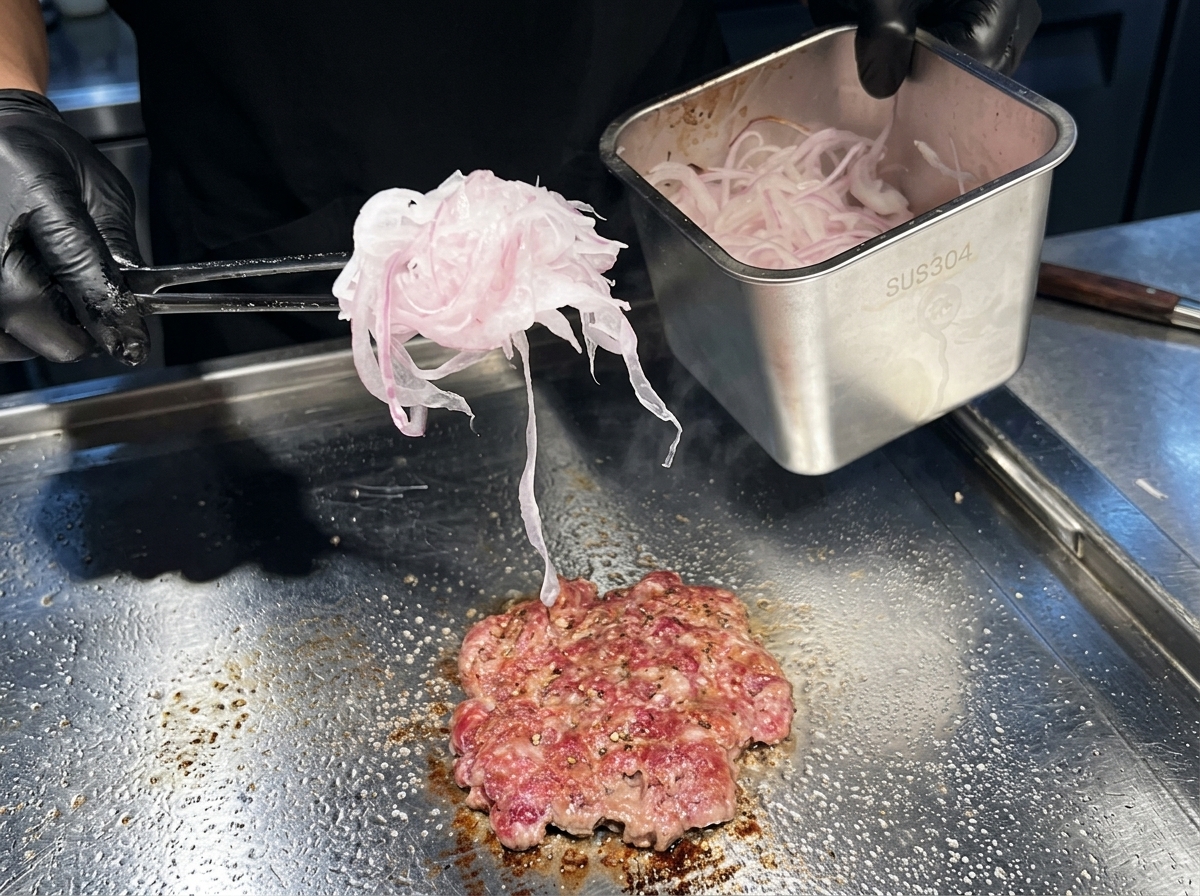 A handful of very thinly sliced red onions being placed on top of a cooking beef patty on a hot griddle.