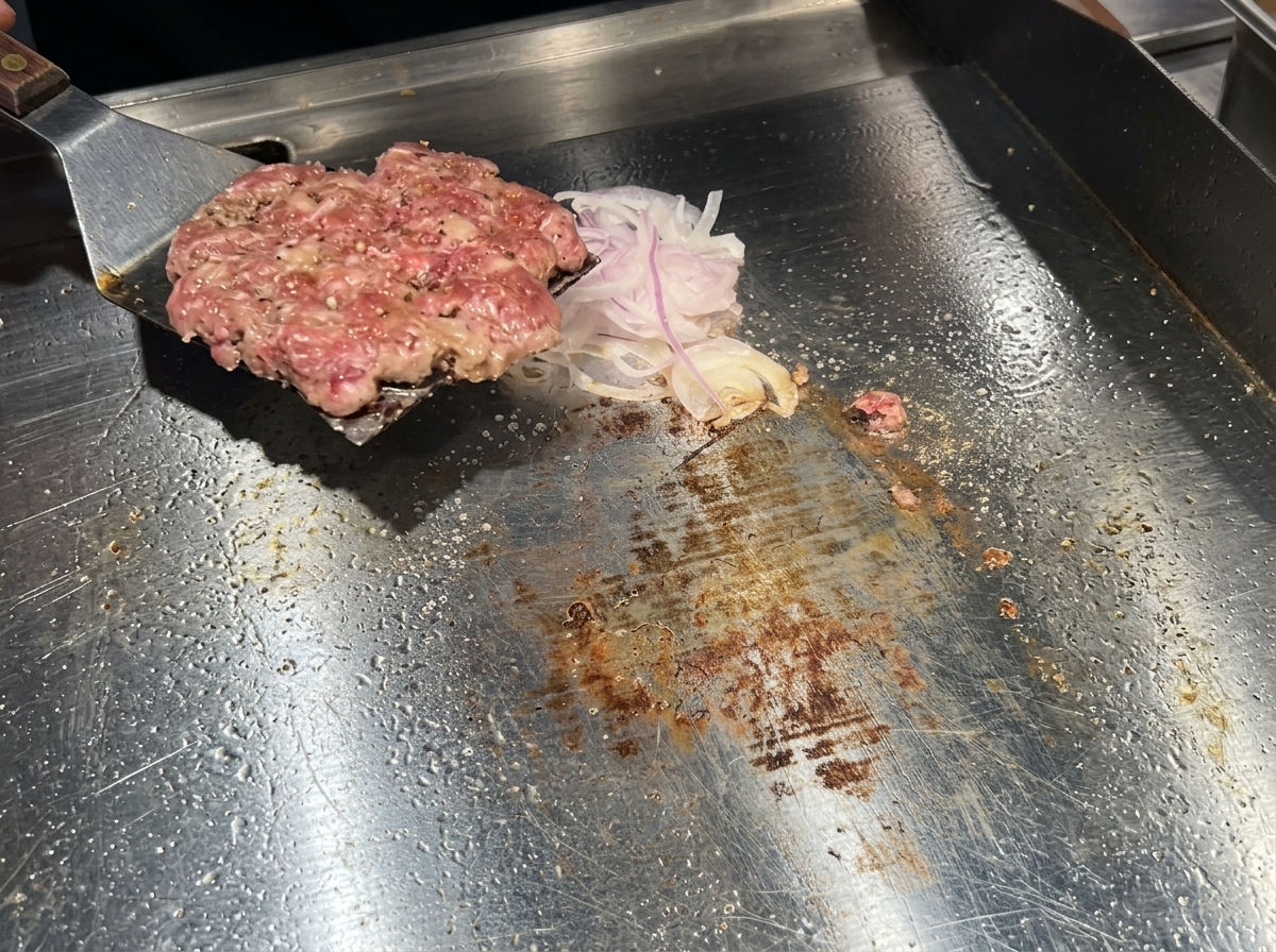 A metal spatula flipping a beautifully browned beef patty over onto a pile of sliced onions on a griddle.