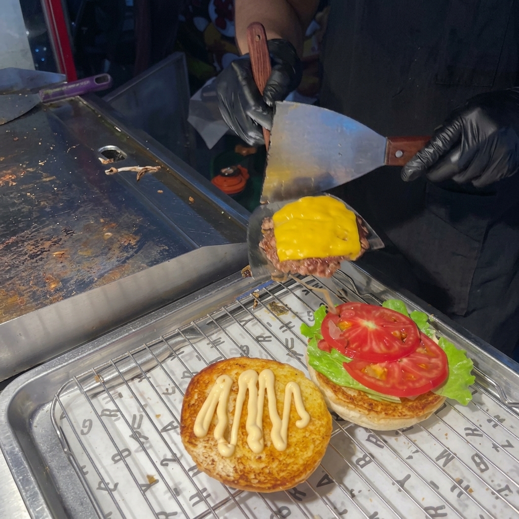 A person using a spatula to place a cooked beef patty with melted cheese onto a burger bun layered with lettuce and tomato.