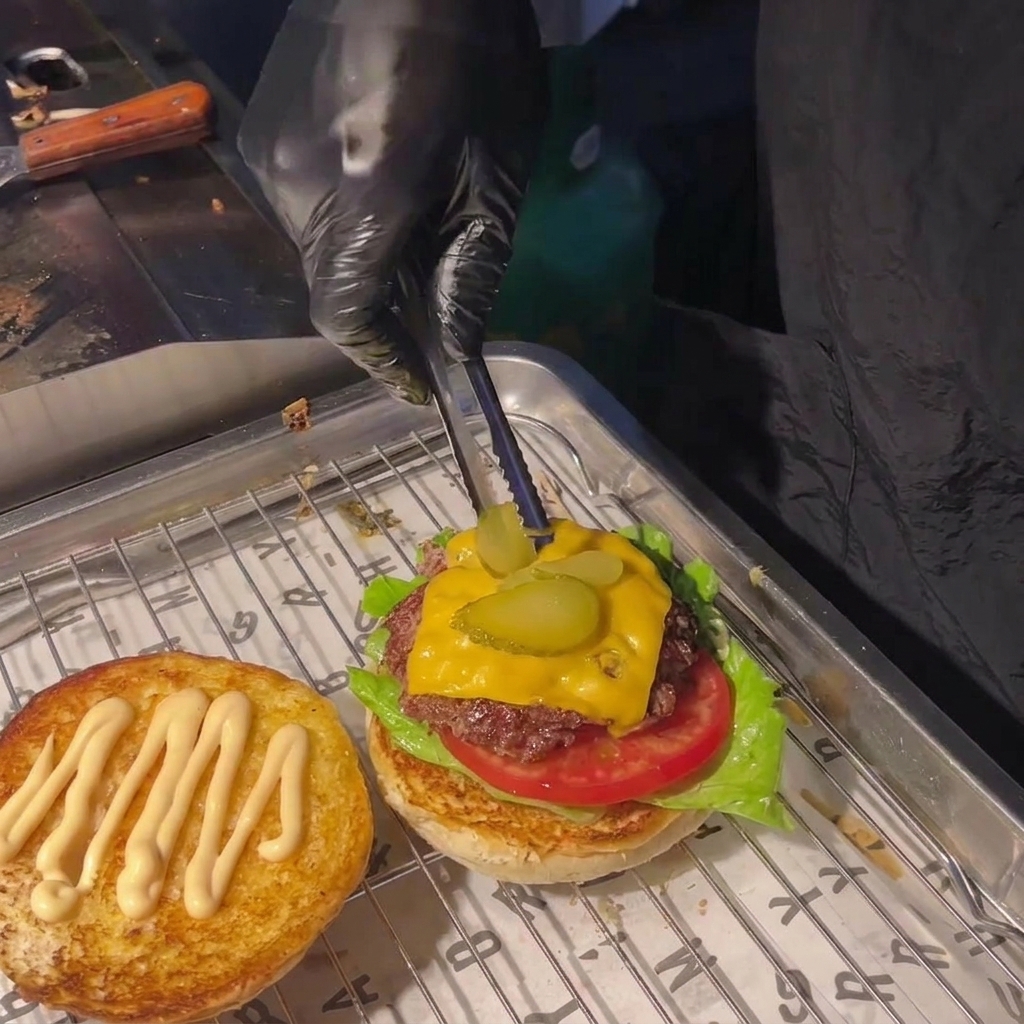 A person using tongs to place two pickle slices on top of the melted cheese on a burger patty.