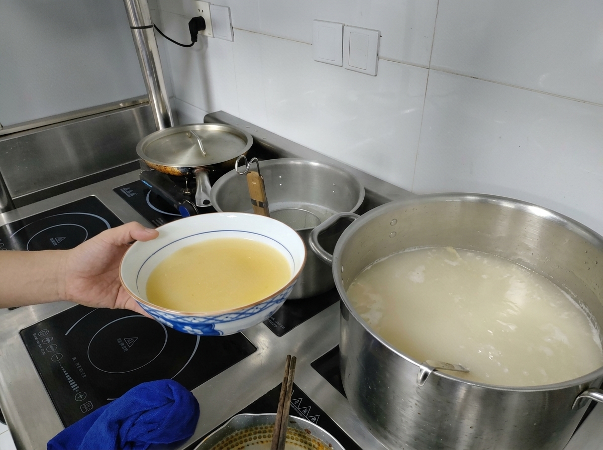 A hand holding a bowl of yellow miso base next to a large pot of boiling milky white broth on a stovetop.