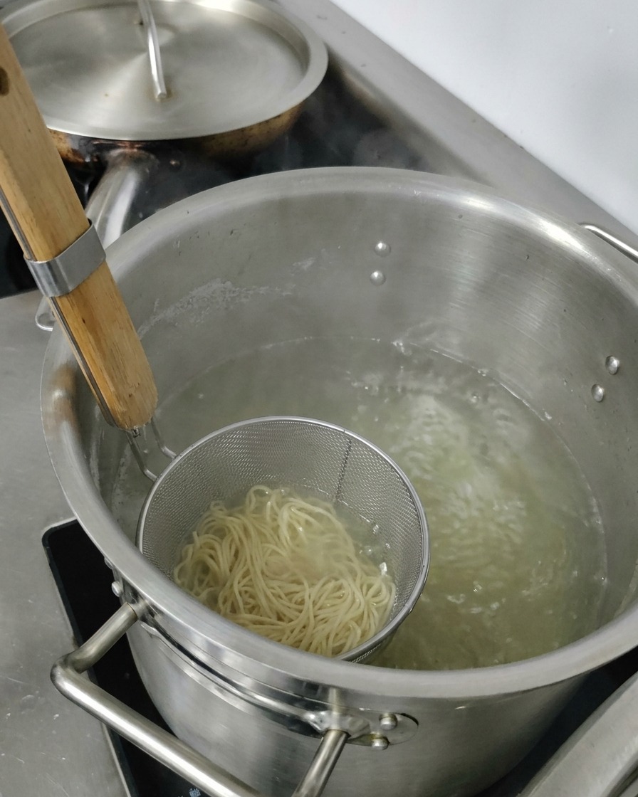 Lifting a metal strainer basket filled with cooked ramen noodles out of a pot of hot water.