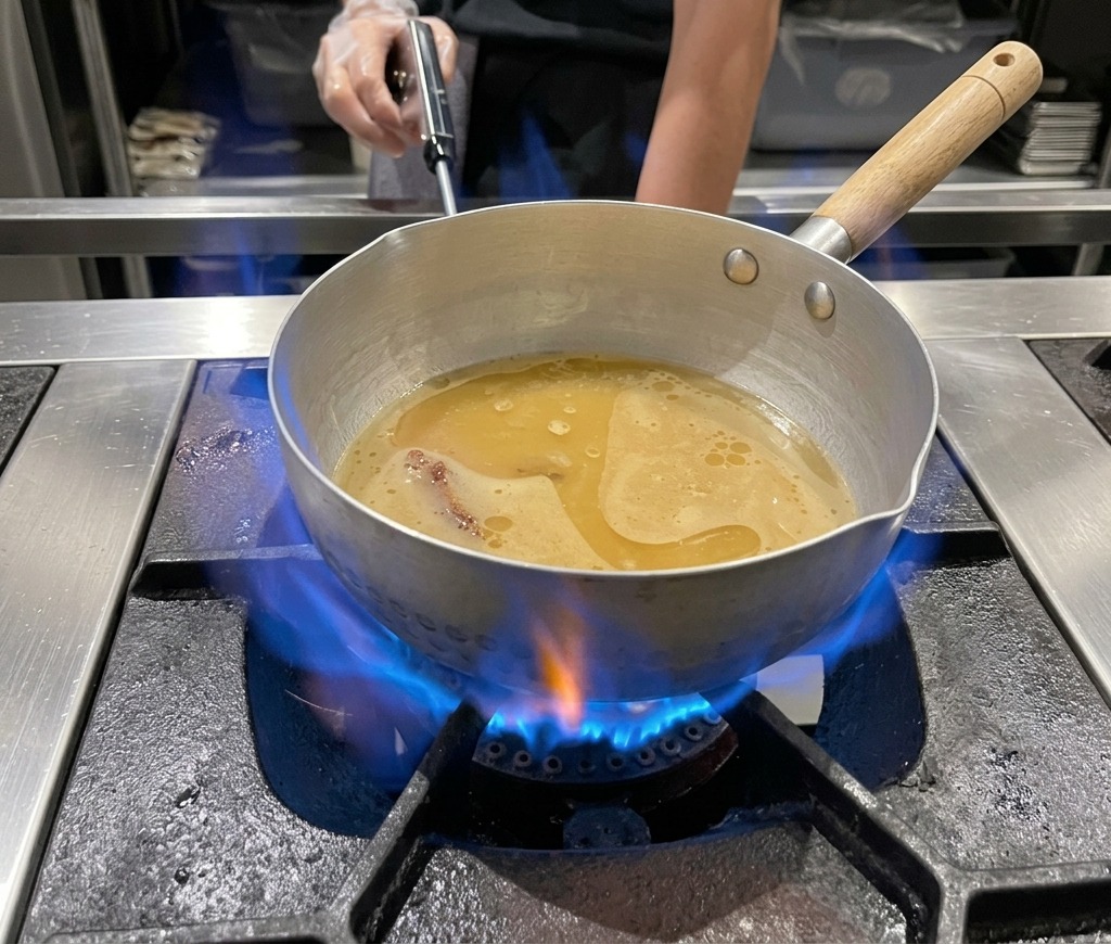 A metal pot of miso ramen broth simmering over a bright blue gas flame on a stovetop.