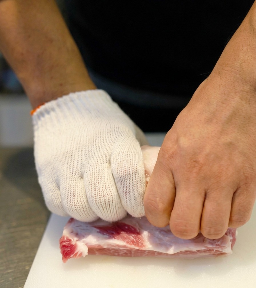 Close-up of hands in white gloves firmly rolling a slab of raw pork belly into a tight log shape.