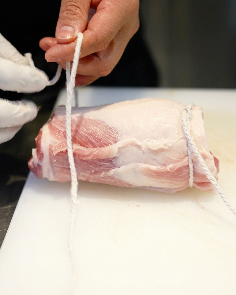 A person using white butchers twine to tie knots around a rolled cylinder of raw pork belly.