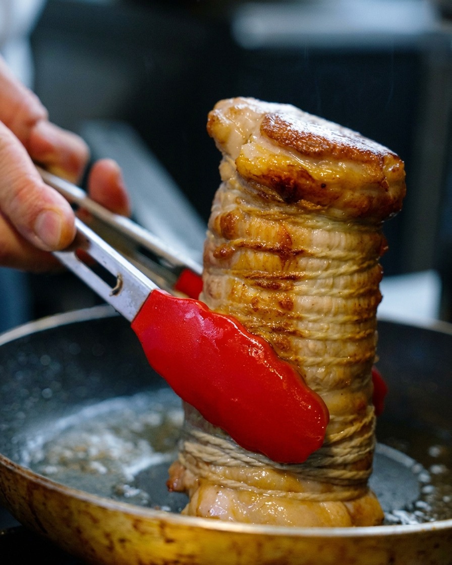 A rolled pork belly tied with twine being held vertically with red-tipped tongs and seared in a pan until golden brown.