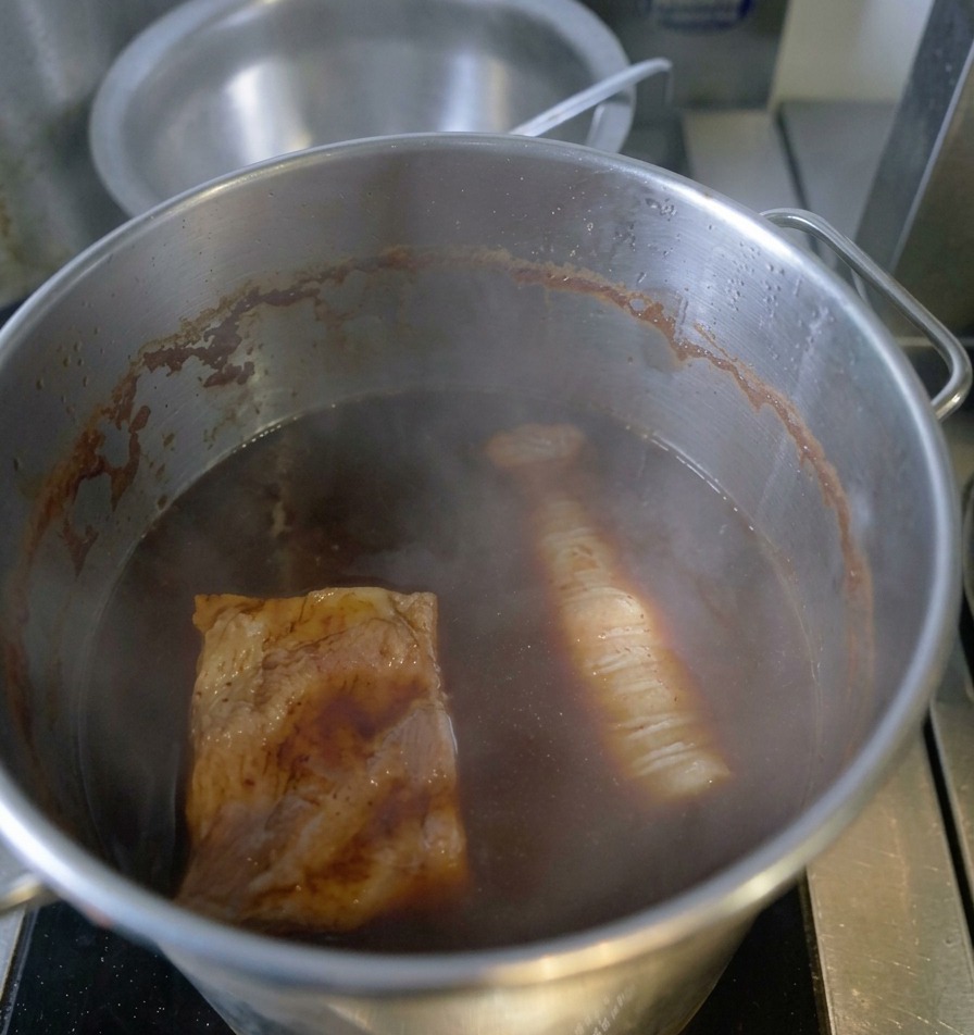 A tied pork roll and another slab of meat simmering in a deep pot of dark, steaming soy-based braising liquid.