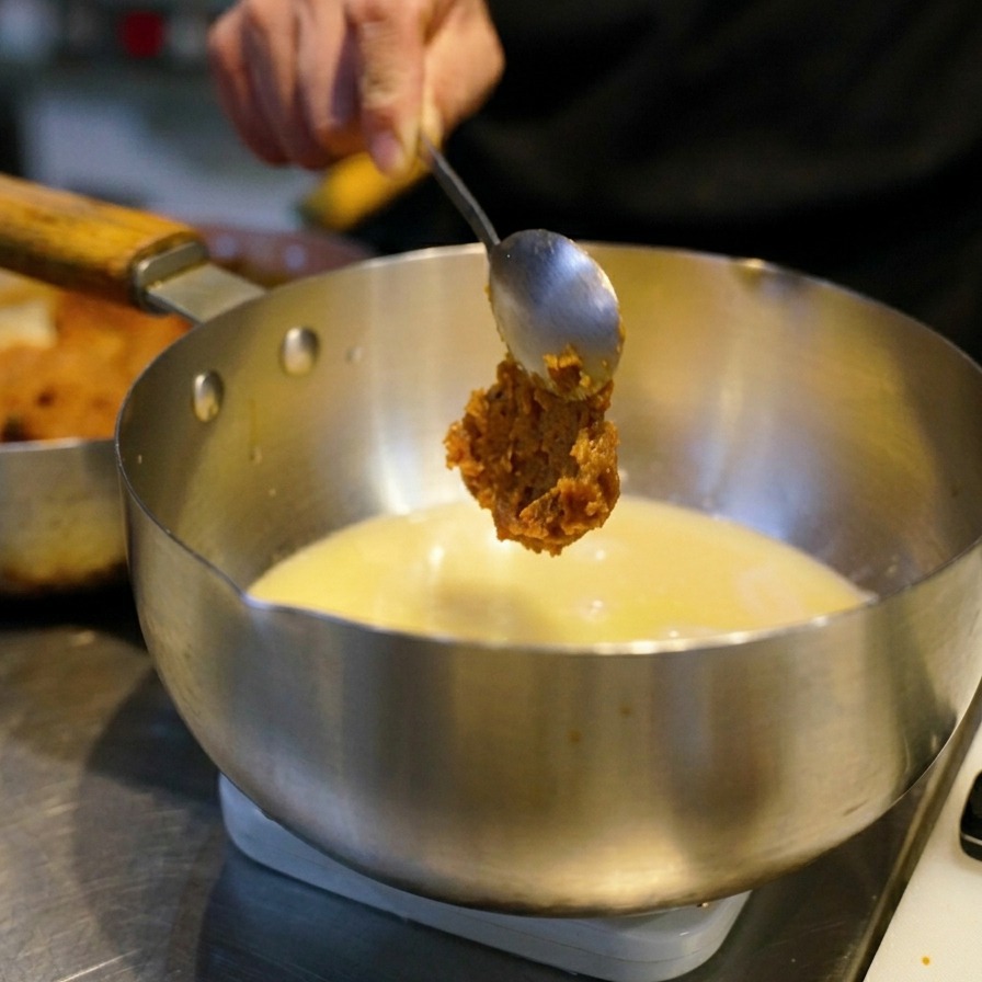 A hand holding a spoon with a large dollop of reddish-brown miso paste over a pot of steaming yellow broth.