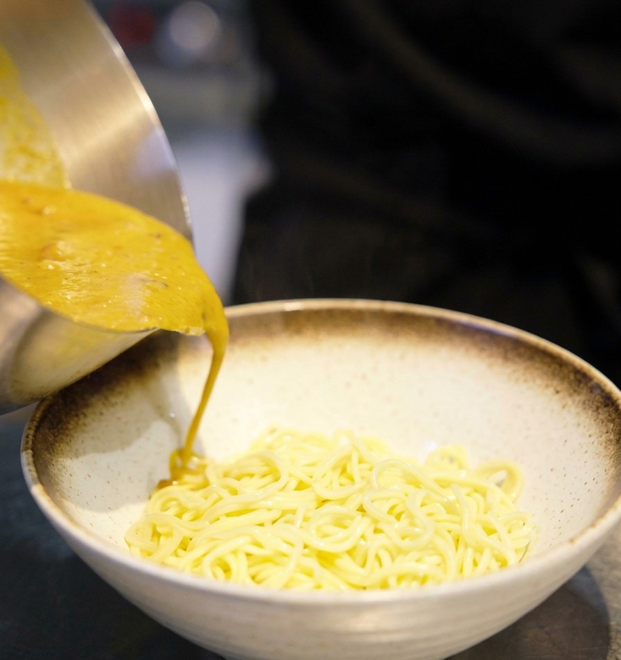 Golden miso broth being poured from a metal saucepan into a white ceramic bowl filled with cooked ramen noodles.