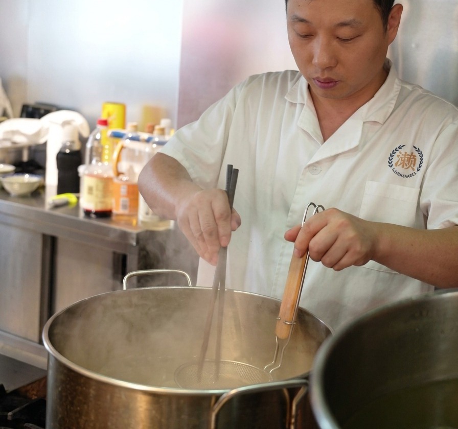 A chef using long wooden chopsticks to stir fresh ramen noodles inside a metal mesh strainer submerged in a large pot of boiling water.