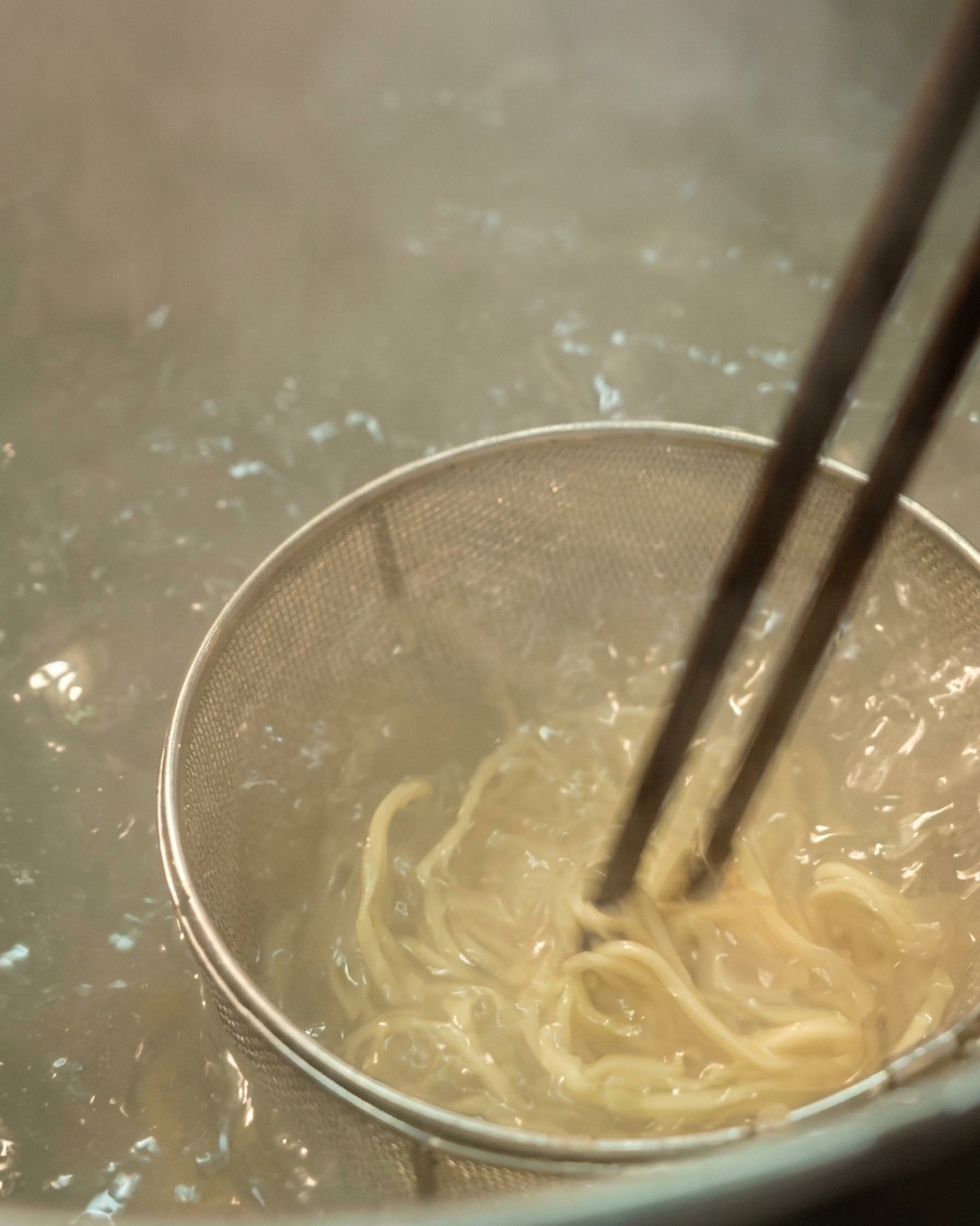 A close up of light colored ramen noodles being stirred with dark chopsticks inside a metal mesh strainer submerged in bubbling boiling water