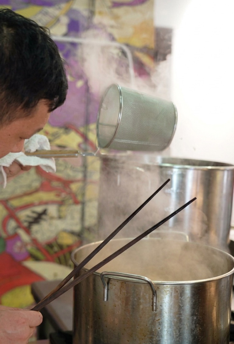 A chef holding a cylindrical mesh noodle strainer over a steaming pot, shaking it to drain water from the ramen noodles inside.
