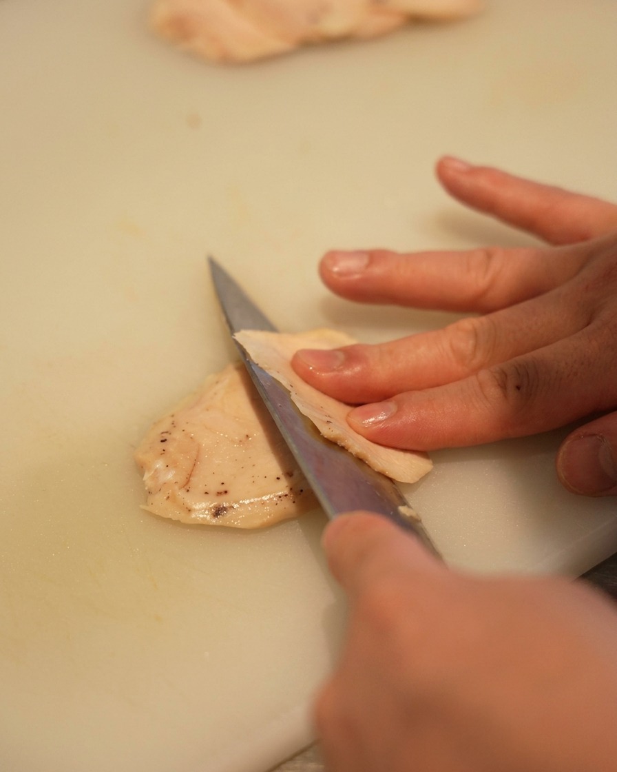Close-up of a hand using a sharp knife to slice a pale chicken breast into thin pieces on a white plastic cutting board.