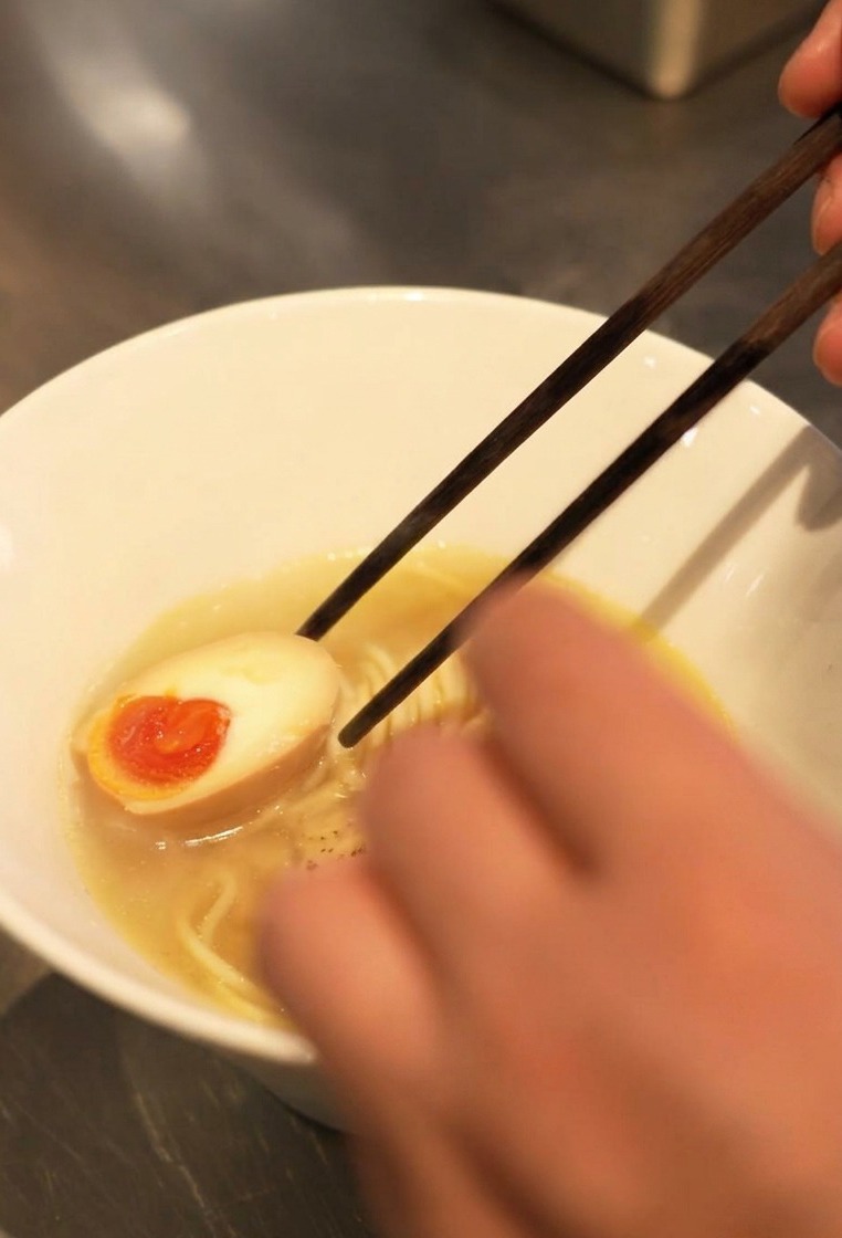 Close-up of chopsticks placing a halved soft-boiled egg with a bright orange runny yolk into a bowl of ramen broth and noodles.