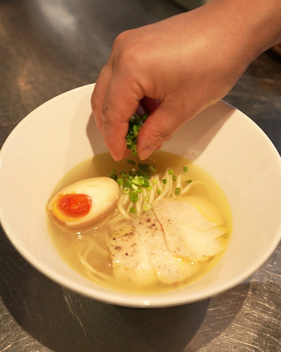 A hand sprinkling freshly chopped green onions over a completed bowl of chicken shio ramen, featuring a soft-boiled egg and sliced chicken.