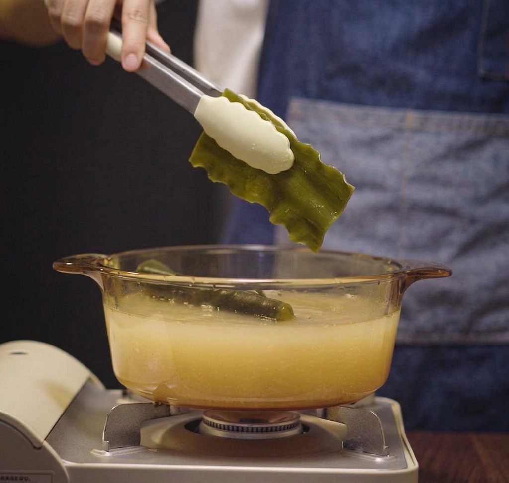 Tongs lowering a folded piece of green kombu into a glass pot containing a cloudy chicken and water mixture.