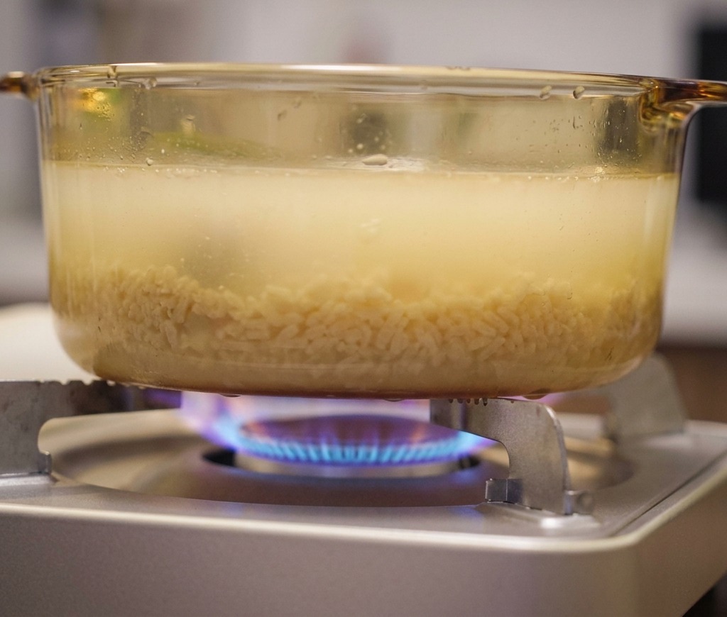A glass pot of cloudy, yellowish ramen broth simmering over a blue gas flame on a portable stove.
