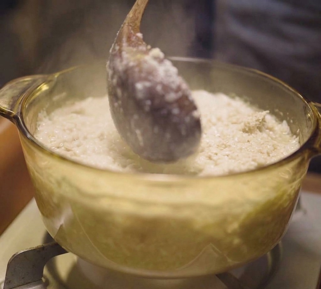 A wooden ladle stirring a pot of simmering white chicken broth with fine cooked meat particles in a clear glass pot.