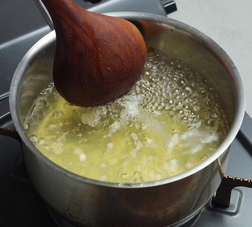 A stainless steel saucepan filled with bubbling, boiling light-colored broth being stirred with a dark wooden spoon.