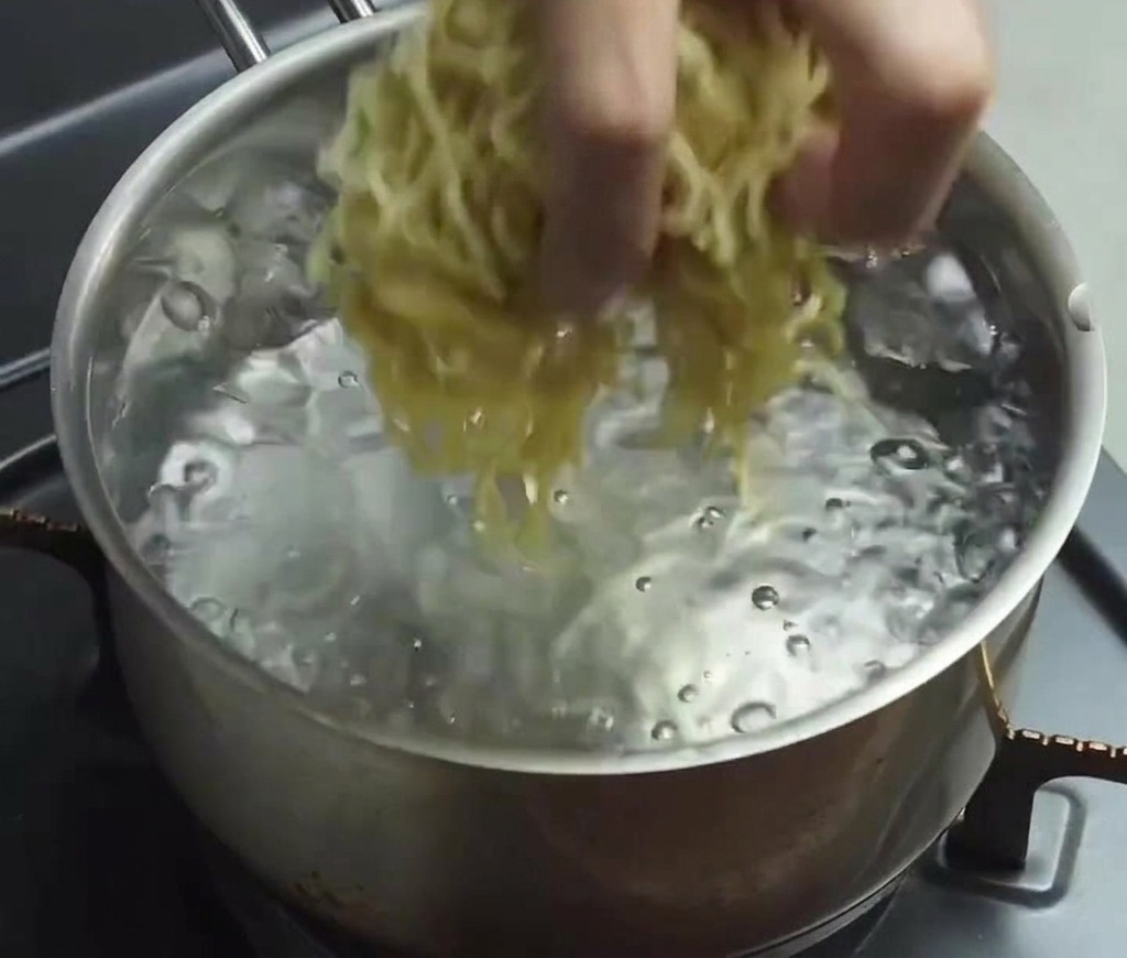 A hand dropping a bundle of thin, wavy yellow fresh ramen noodles into a pot of clear, rapidly boiling water.