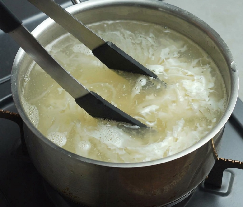 Black-tipped tongs stirring yellow noodles in a pot of boiling water on a stove burner.