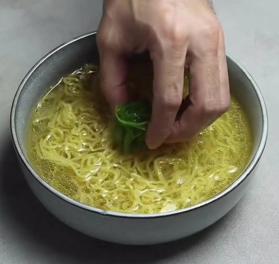 A hand placing a cluster of thin green onion shreds onto ramen noodles in a bowl of clear yellow broth.