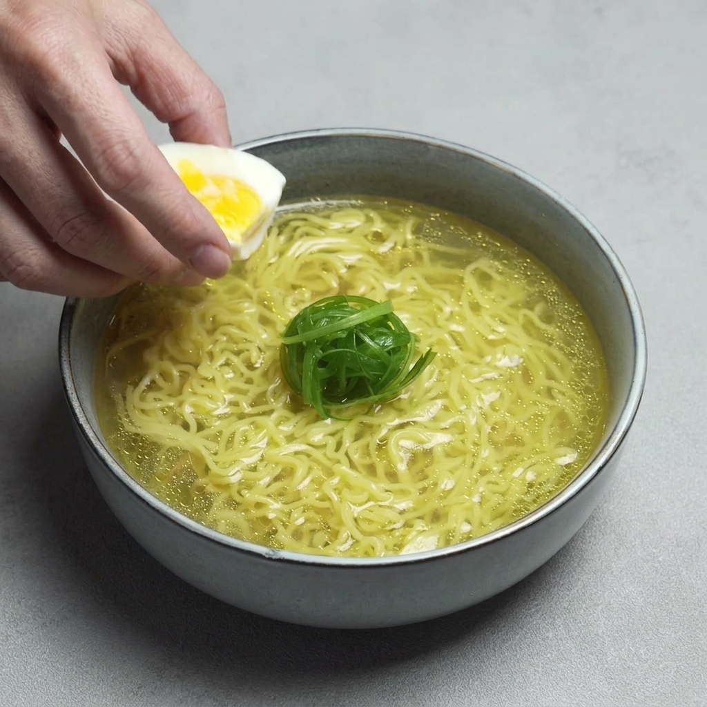 A hand placing a half of a soft-boiled egg with a bright yellow yolk into a bowl of ramen already garnished with a curl of green onions.