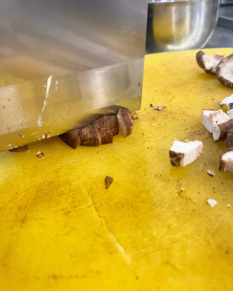 A large kitchen knife dicing brown shiitake mushroom caps into small cubes on a bright yellow cutting board.