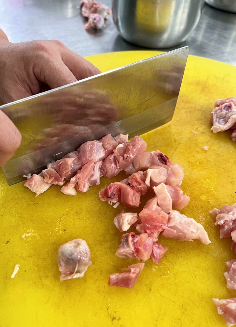 A person using a large metal cleaver to dice raw chicken meat on a bright yellow plastic cutting board.