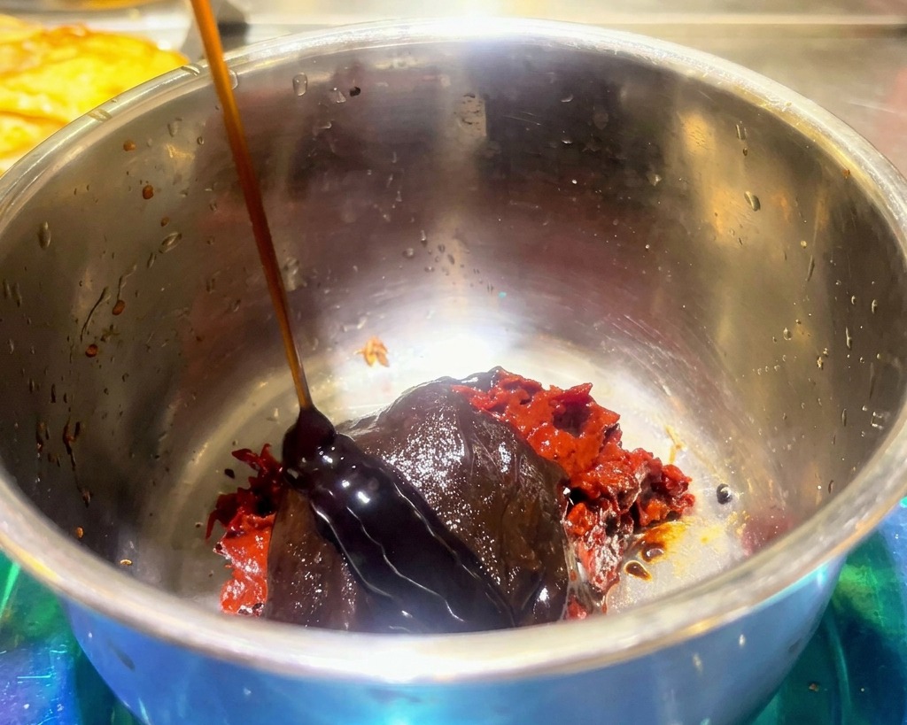 Thick dark sauce being poured over red spicy bean paste and hot pot seasoning in a stainless steel bowl.