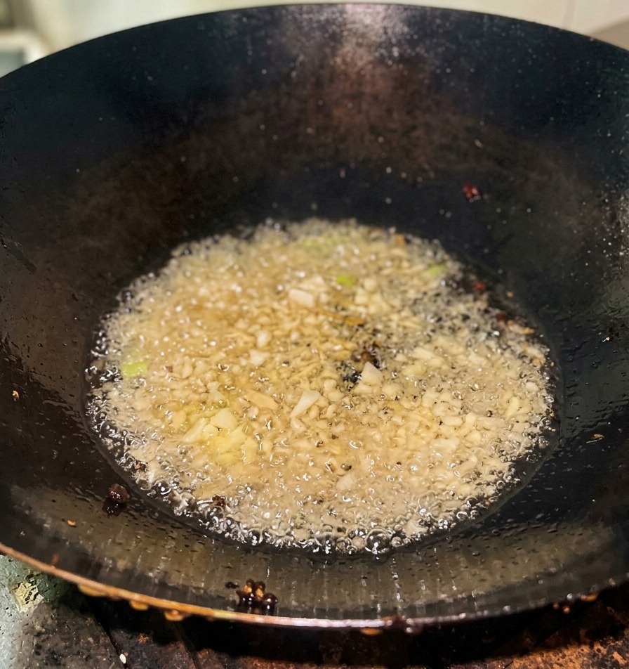 Chopped green onions sizzling and bubbling in clear hot oil at the bottom of a dark iron wok.