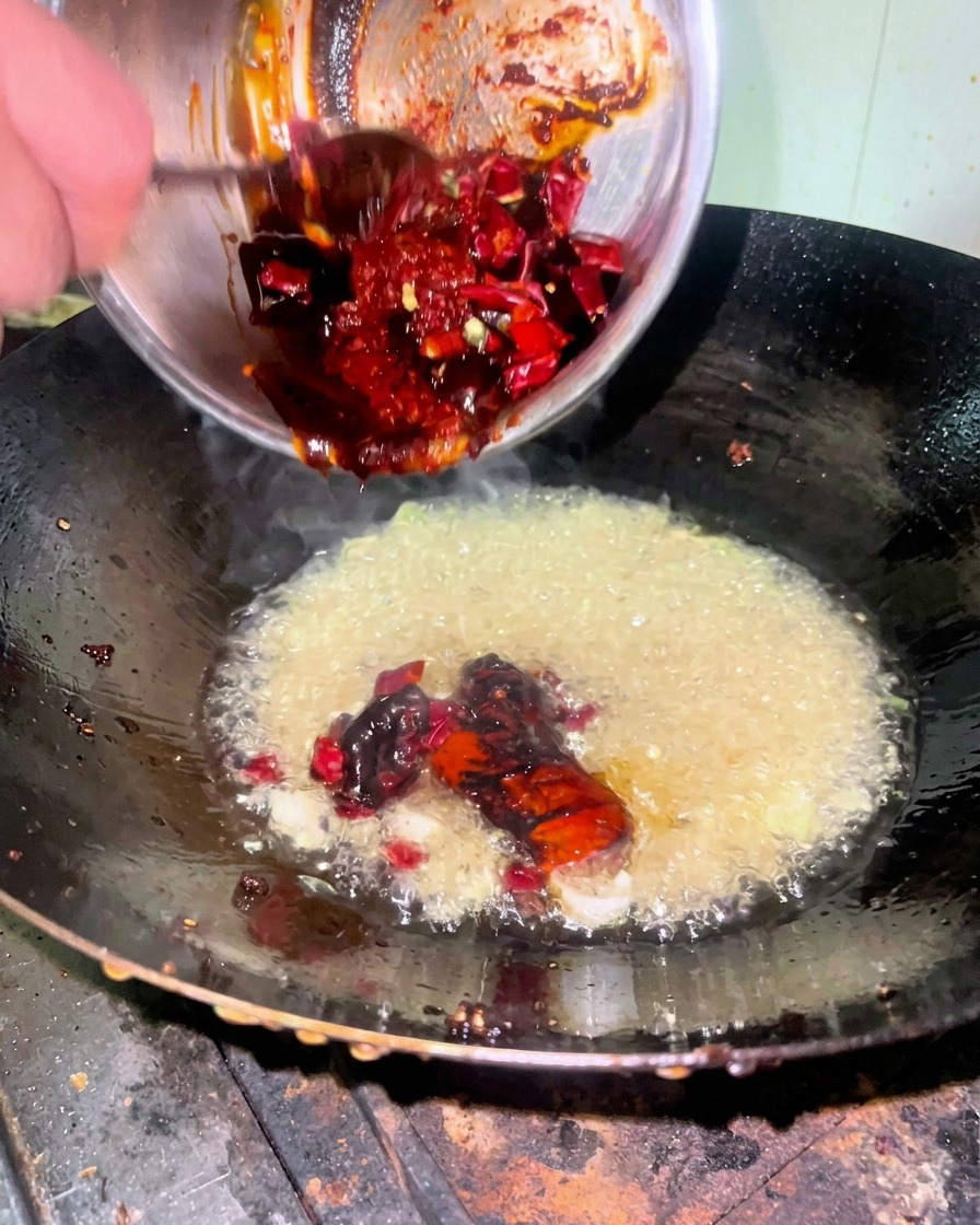 A thick, dark red spicy sauce mixture being scraped from a metal bowl into a wok containing hot oil and aromatics.