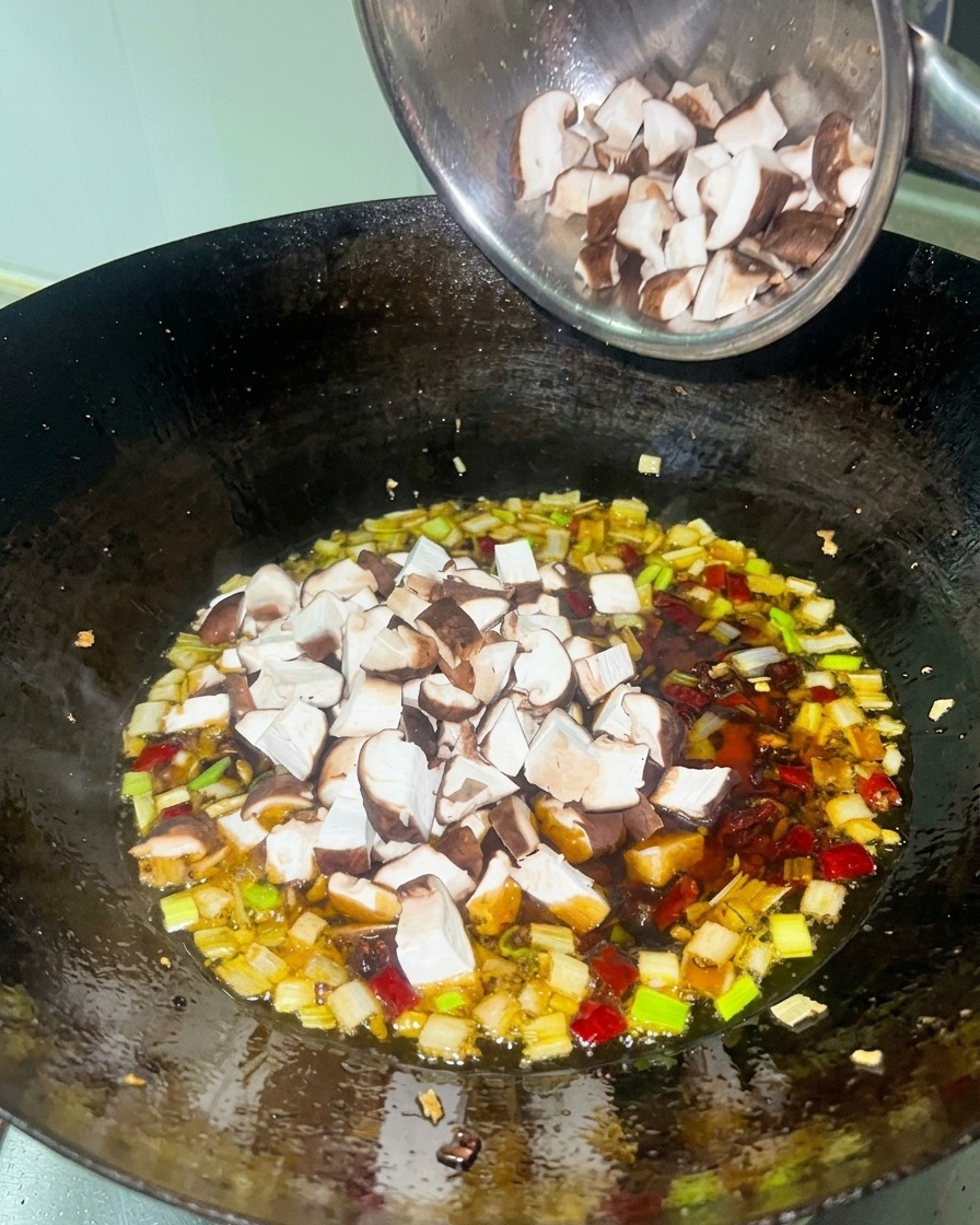 Diced white chicken meat and brown shiitake mushrooms being added to a rich, red simmering sauce in a black wok.