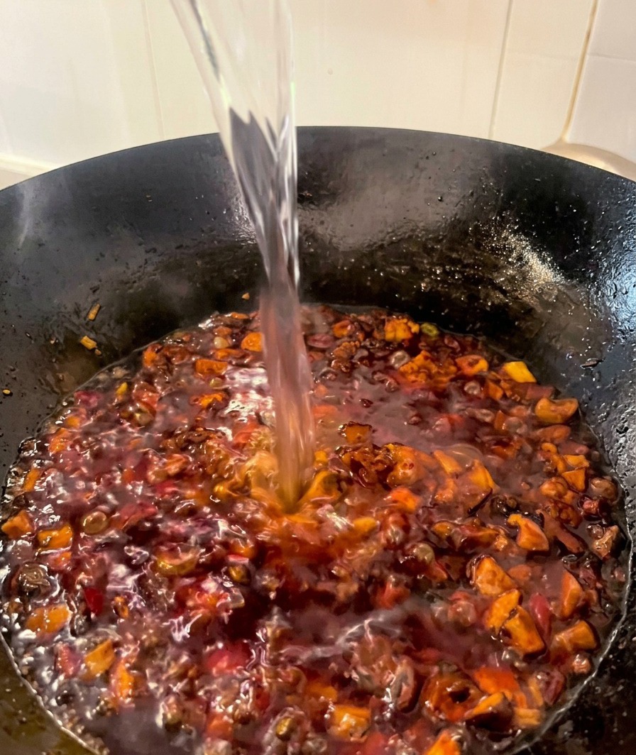 A steady stream of clear water being poured into a wok filled with a dark red, simmering chicken and mushroom sauce.