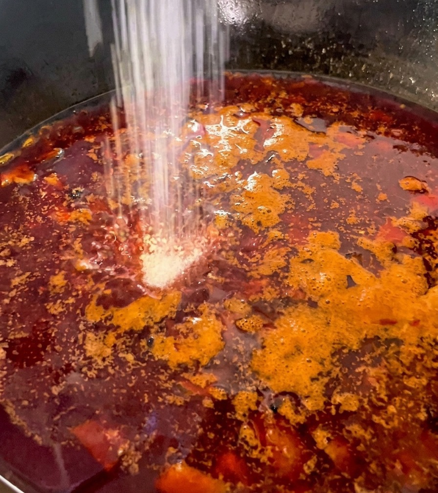 White seasoning powder being sprinkled into a deep red, bubbling broth containing diced mushrooms and chicken.