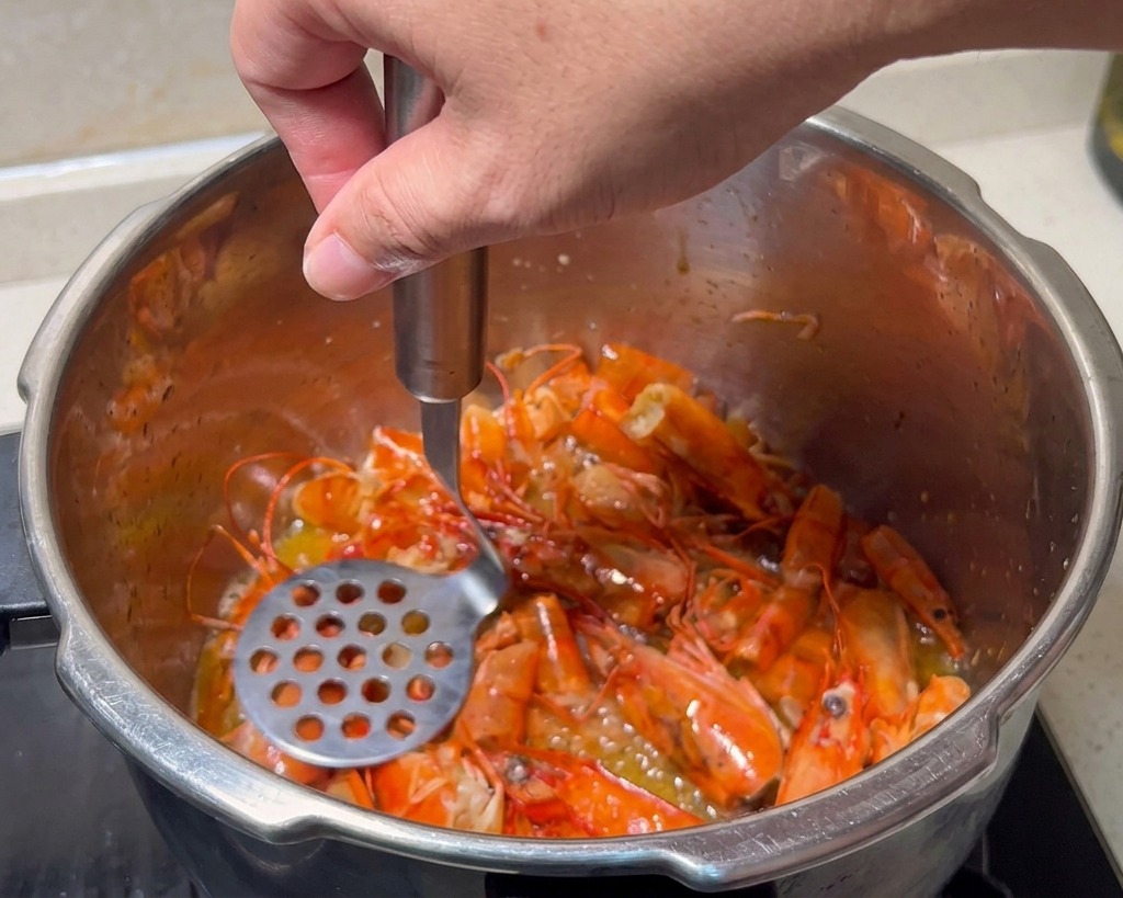 A metal potato masher being used to crush bright red fried shrimp heads in a stainless steel pot.