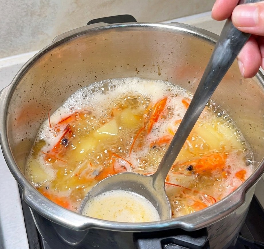 A metal ladle stirring a bubbling, orange-tinted shrimp broth with visible shrimp heads and melting butter.