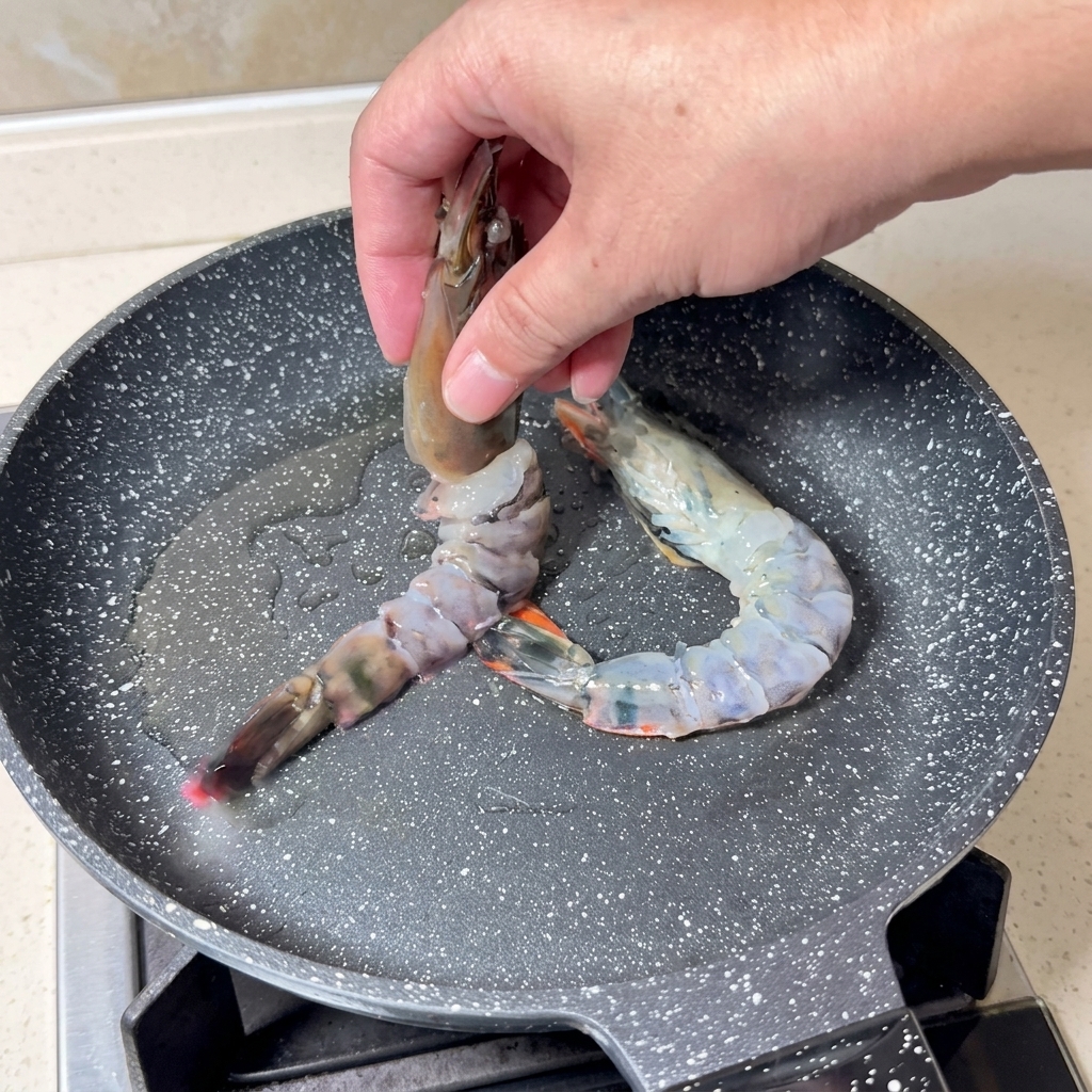 A hand placing large, butterflied raw shrimp into a hot, speckled grey frying pan with shimmering oil.