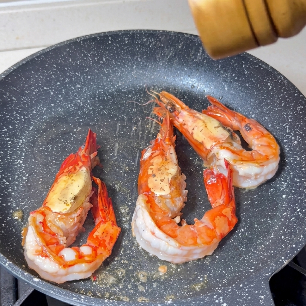 Salt and pepper being ground over three large shrimp cooking in a pan, showing bright orange shells and white cooked flesh.