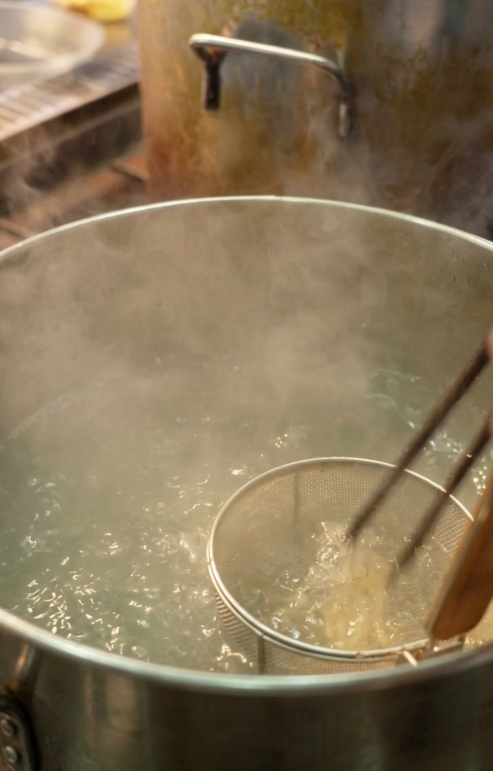 A chef using a mesh noodle strainer to dunk thin white ramen noodles into a large pot of steaming, boiling water.