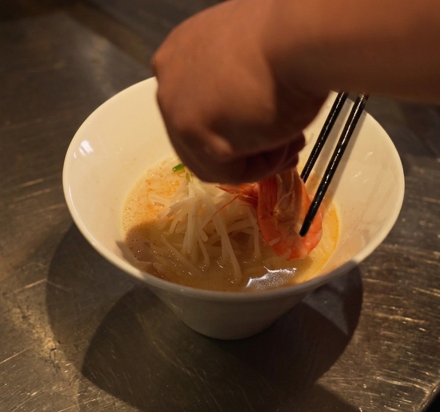 A hand using chopsticks to place a cooked, curved shrimp onto a bed of noodles and bean sprouts in a white bowl.
