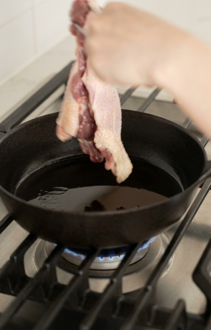 A hand using tongs to place a raw duck leg skin side down into a hot oiled cast iron skillet