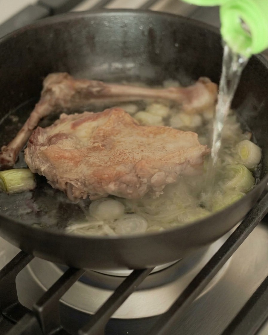 Clear sake being poured from a bottle into a steaming skillet containing seared duck and sliced leeks.