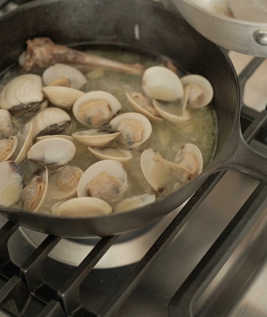 Clams being lifted from a cast-iron skillet containing a duck bone and savory broth using metal tongs.