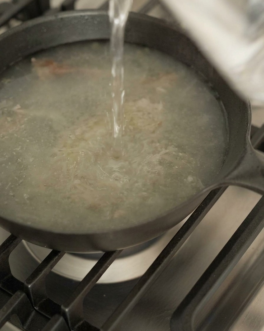 Water being poured from a height into a dark skillet containing meat juices and aromatics on a gas stove.