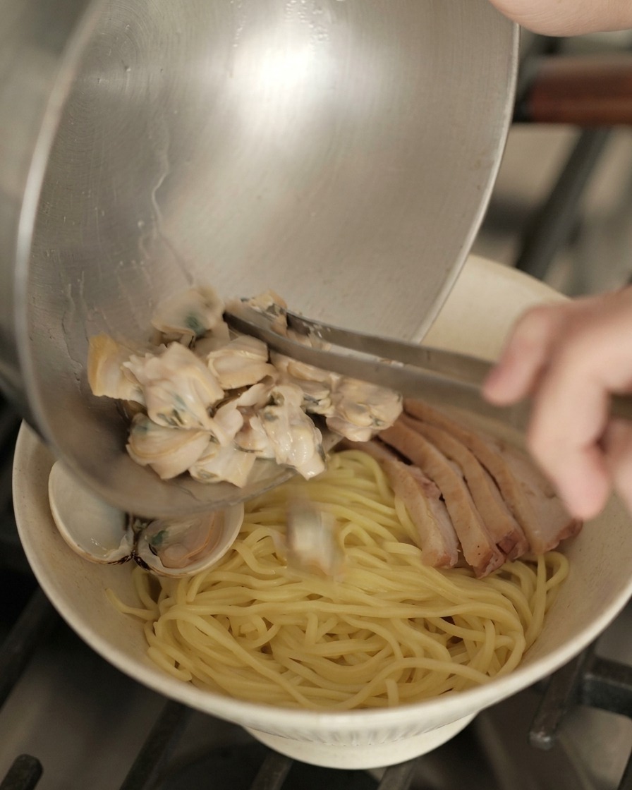 Pouring cooked, opened clams from a metal bowl onto a prepared bowl of ramen noodles and duck.