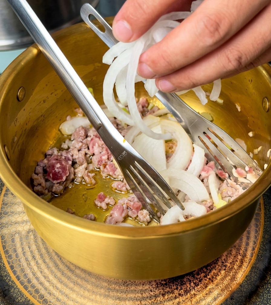 Hand adding sliced white onions into a golden pot containing sautéed ground pork and aromatics.