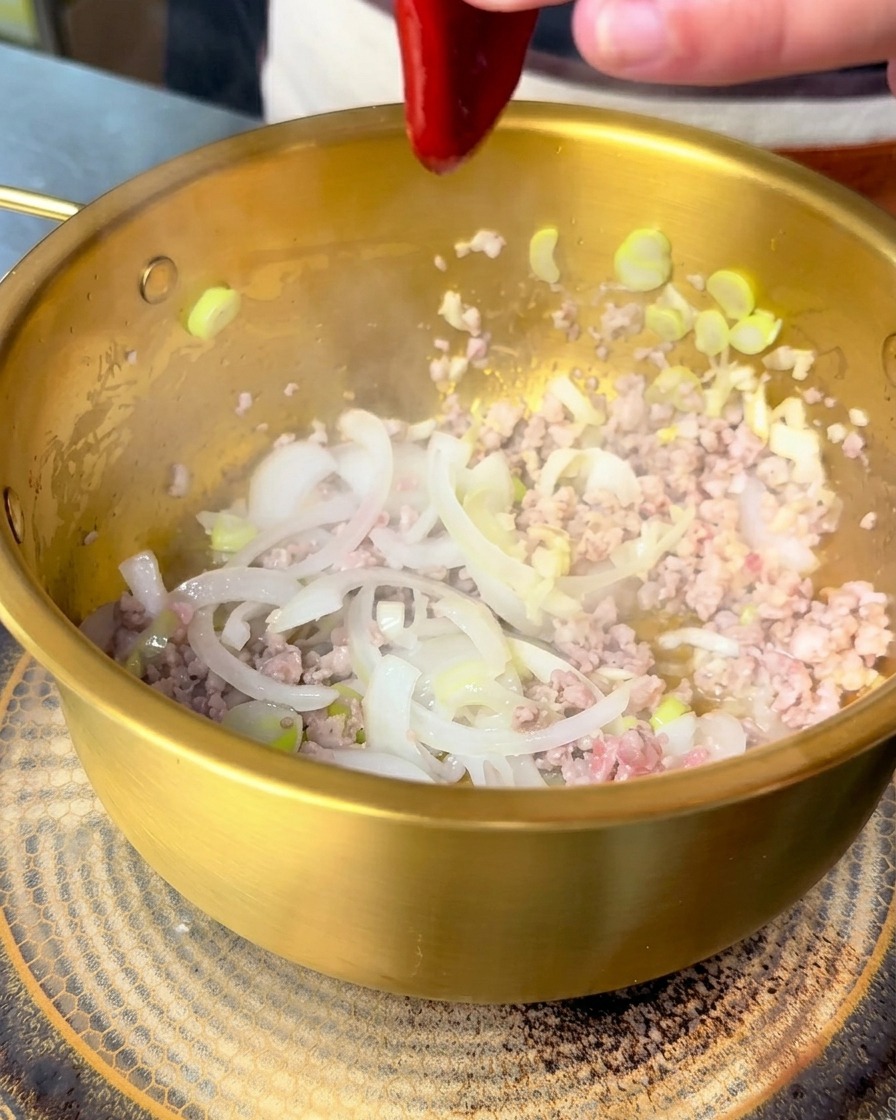 Red Korean bibimbap sauce being squeezed into a golden pot containing stir-fried pork and onions.